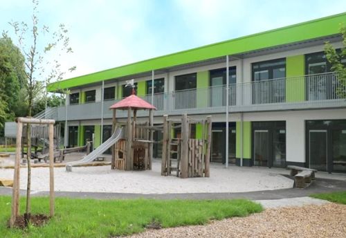 Die Kita Spatzennest von außen betrachtet und der Spielplatz mit Kletterturm, Rutsche, Wippe und weiteren Spielgeräten.